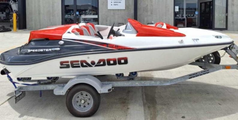 Puede incluir: Un barco Sea Doo Speedster 200 blanco y negro sobre un remolque. El barco tiene detalles y asientos rojos. El nombre del barco, "SEA DOO 200", se muestra en letras negras grandes en el lateral.