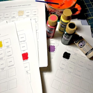 May include: An assortment of art supplies, including paint swatch cards, paint bottles, and a decorative orange bowl with paint. The paint bottles are labeled "Americana" and "DecoArt". The swatch cards display various paint colors.