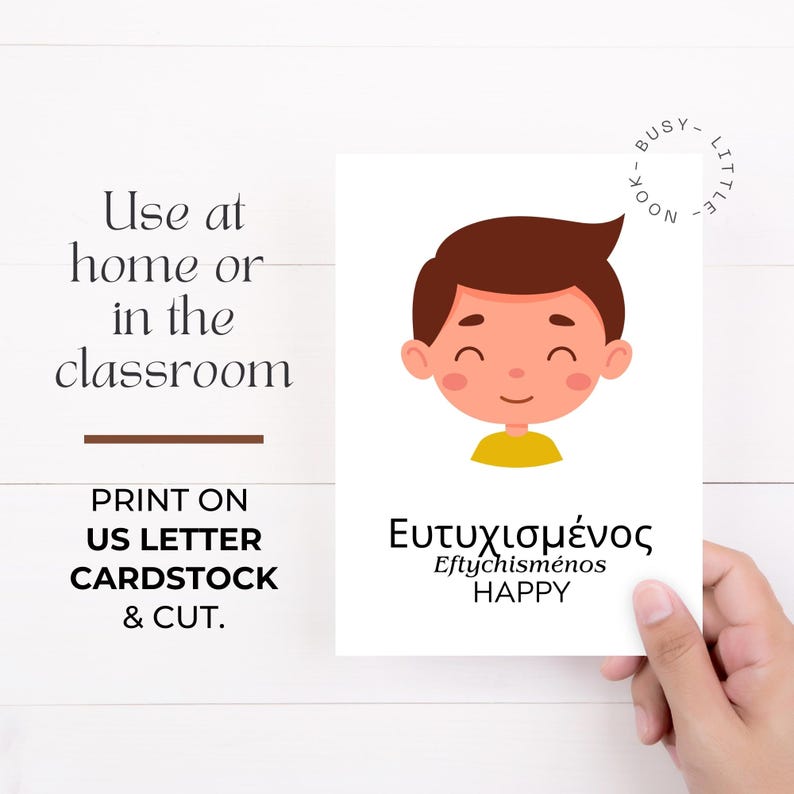 May include: A white card with a cartoon illustration of a smiling boy with brown hair and a yellow shirt. The card has the Greek word "&Epsilon;&upsilon;&tau;&upsilon;&chi;&iota;&sigma;&mu;έ&nu;&omicron;&sigmaf;" and the English word "HAPPY" printed on it. Text on the card reads "Use at home or in the classroom."