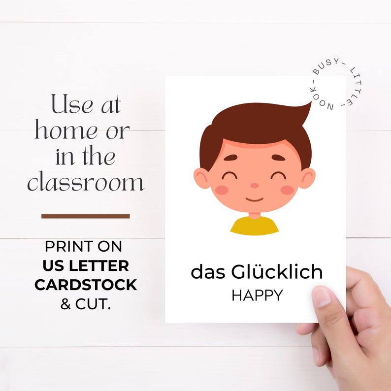 Puede incluir: Una tarjeta blanca con una ilustraci&oacute;n de dibujos animados de un ni&ntilde;o sonriente con cabello casta&ntilde;o y una camisa amarilla. La tarjeta tiene la palabra alemana "das Gl&uuml;cklich" y la palabra inglesa "HAPPY". El texto a la izquierda dice "Usar en casa o en el aula."