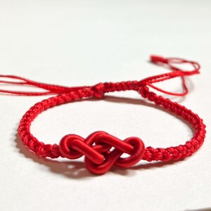 Puede incluir: Una pulsera roja brillante con un diseño central de nudo infinito. La pulsera está hecha de cordón rojo tejido, con lazos ajustables para un ajuste cómodo. El nudo es un punto focal prominente.