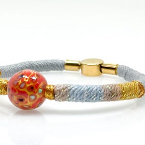 May include: A colorful bracelet featuring a red and gold patterned bead. The bracelet has a gray, gold, and blue woven design with a gold magnetic clasp. A stylish accessory.