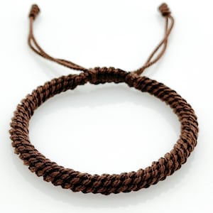 May include: A brown braided bracelet with adjustable cord ties. The bracelet is made of tightly woven, dark brown material, creating a textured appearance. The adjustable ties allow for a customized fit.