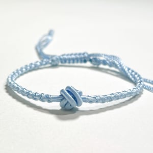 May include: A light blue macrame bracelet with a decorative knot in the center. The bracelet is made of woven cord and has adjustable sliding knots. The bracelet is shown on a white background.