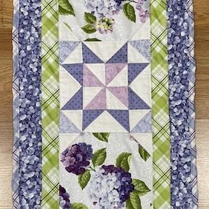 May include: A rectangular table runner featuring a patchwork design. The center panel has star patterns in white, lavender, and purple. Hydrangea floral patterns are interspersed. The border is a blue floral print and green plaid.