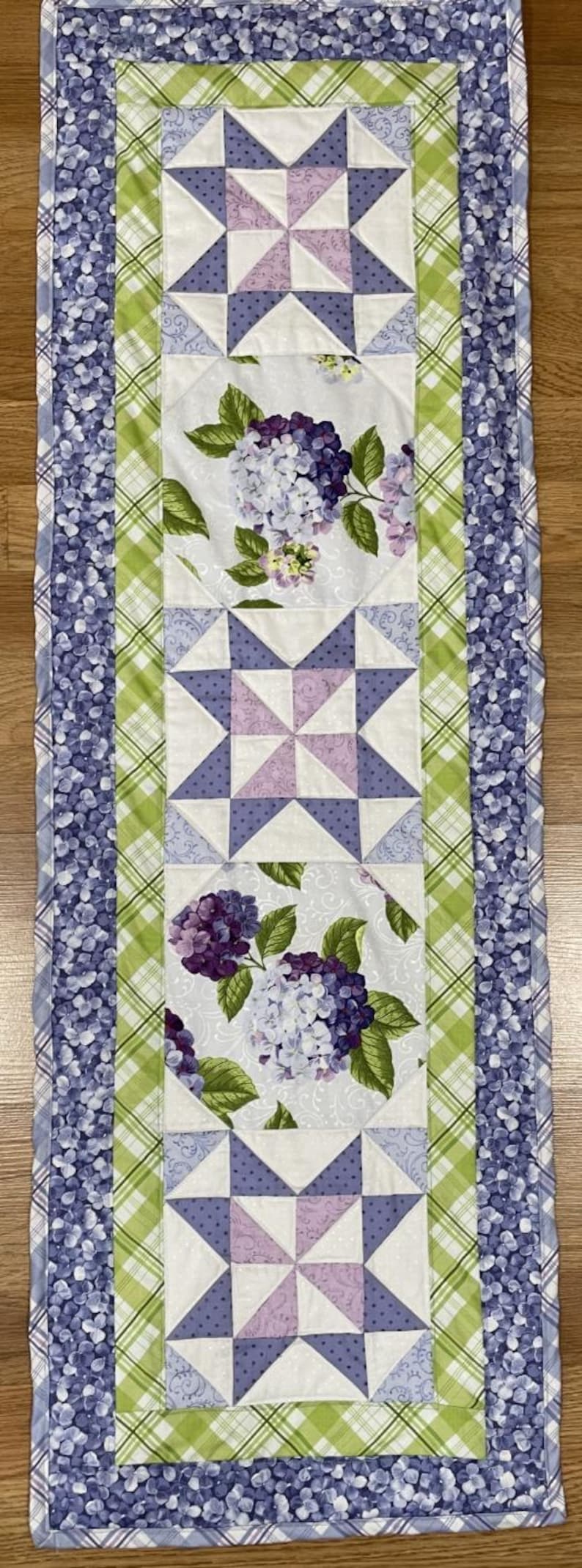May include: A rectangular table runner featuring a patchwork design. The center panel has star patterns in white, lavender, and purple. Hydrangea floral patterns are interspersed. The border is a blue floral print and green plaid.