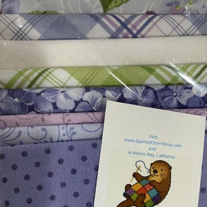 May include: A stack of fabric bundles in various pastel colors, including lavender, white, and green, with floral and plaid patterns. A card with an otter illustration and the text "The Quilted Otter" is visible.