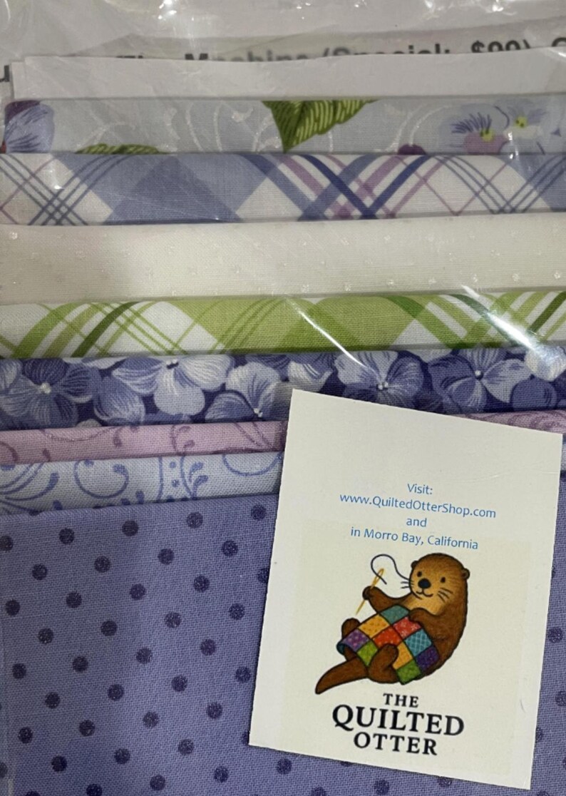 May include: A stack of fabric bundles in various pastel colors, including lavender, white, and green, with floral and plaid patterns. A card with an otter illustration and the text "The Quilted Otter" is visible.