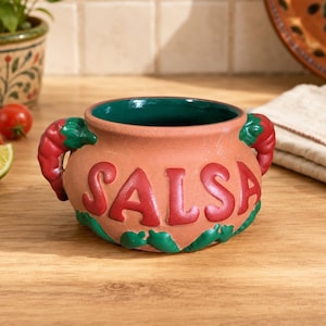 May include: A terracotta salsa bowl with a dark green interior and two red chilli pepper handles. The word SALSA is embossed in large red letters on the front, and the base is decorated with small green chilli pepper motifs in relief.