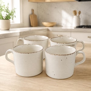 May include: A set of four off-white ceramic mugs with a speckled finish and a brown rim. Each mug features a wide, cylindrical body and a sturdy side handle. The mugs are arranged in a group on a light-colored wooden surface, showcasing a consistent design.