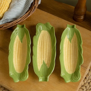 May include: Three ceramic corn-shaped serving dishes with a green husk design and a yellow corn interior. The dishes are arranged on a wooden cutting board. A basket with corn on the cob and a wooden pepper grinder are in the background.