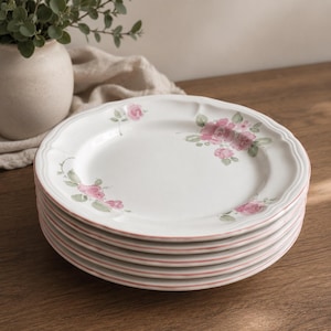 May include: A stack of white ceramic plates featuring a delicate floral pattern of pink roses and green leaves. Each plate has a scalloped edge with a thin pink painted rim. The design is consistent across the set, showcasing soft pastel colours.