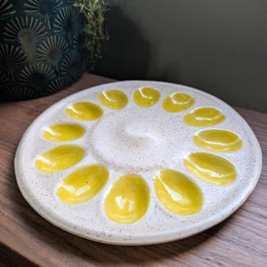 May include: A speckled white ceramic egg plate with 12 yellow egg-shaped indentations. The plate is round and sits on a wooden surface. A dark teal decorative pot with a plant is in the background.