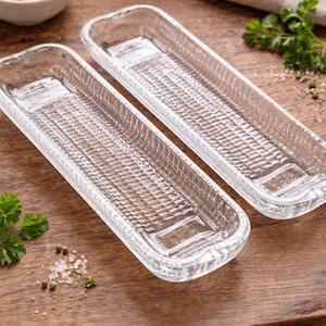 May include: Two clear, rectangular glass serving dishes with a textured pattern. The dishes are on a wooden surface, with a small bowl of butter and sprigs of parsley in the background. The dishes are designed for serving food.