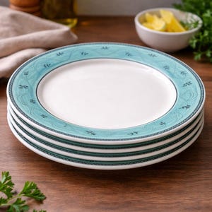 May include: Four white ceramic plates with a teal border and decorative pattern, stacked on a wooden surface. The plates are designed for dining. The plates have a simple, elegant design. The plates are stacked on a wooden surface.