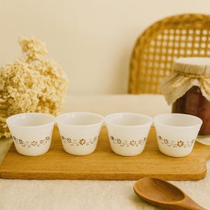 May include: Four white ceramic bowls with a floral design in a warm brown hue are arranged on a wooden cutting board. The bowls are small and have a slightly flared shape. A wooden spoon rests nearby.