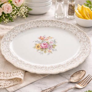 May include: White oval serving platter with a floral centre and gold filigree edging. The platter has a scalloped edge. Silverware and a linen napkin are next to the platter. The platter measures approximately 30 cm.