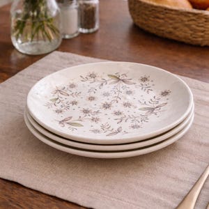 Peut inclure: Trois assiettes en céramique blanche empilées avec un motif floral. Les assiettes présentent un motif délicat de petites fleurs et de feuilles dans des tons verts, bruns et bleus discrets. Les assiettes sont posées sur un set de table en lin beige.