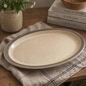 May include: An oval ceramic serving platter with a speckled cream surface and a textured, grey-brown border. The platter sits on a linen cloth, with books and a wooden bowl in the background. The books have the titles "THIS IS HOME" and "KINFOLK".