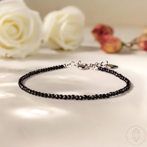 Könnte beinhalten: Ein zartes Armband mit schwarzen Perlen und einem silbernen Verschluss. Das Armband besteht aus kleinen, runden, schwarzen Perlen. Der Hintergrund zeigt weiße Rosen und andere Blumen, was eine weiche, elegante Ästhetik erzeugt.