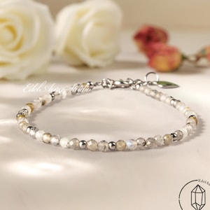 Könnte beinhalten: Ein filigranes Armband mit kleinen, facettierten Edelsteinen in Weiß, Grau und Gold, durchsetzt mit silberfarbenen Perlen. Das Armband wird auf einer hellen Oberfläche präsentiert, mit unscharfen Rosen im Hintergrund. Es hat einen silbernen Verschluss.