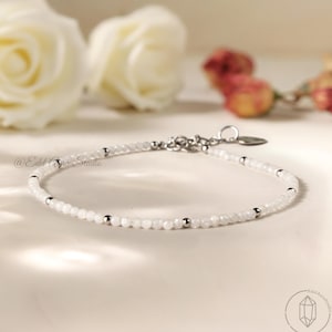 Può includere: Un delicato braccialetto di perline bianche con dettagli argentati. Il braccialetto presenta piccole perline bianche rotonde intervallate da perline argentate e una chiusura argentata. Lo sfondo include rose bianche e altri fiori.