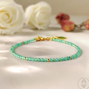 May include: A fine bracelet with small, round, light green beads and gold-coloured accents. The bracelet is strung on a thin cord and has a gold clasp. The background includes white roses and the text "@Edel Aura Studio".