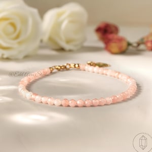 Può includere: Un delicato braccialetto con piccole perline rosa sfaccettate. Il braccialetto ha una chiusura color oro ed è esposto con rose bianche e altri fiori. Il testo "@Edel Aura Studio" è visibile.