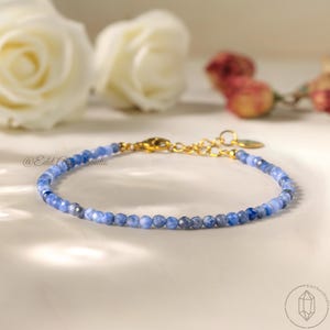 Può includere: Delicato braccialetto con piccole perline blu sfaccettate e chiusura dorata. Lo sfondo presenta rose bianche e fiori secchi. Il braccialetto è esposto su una superficie bianca. Un gioiello elegante e raffinato.