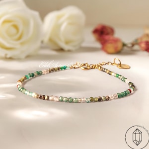 Könnte beinhalten: Zartes Perlenarmband mit einer Mischung aus grünen, braunen und rosa Edelsteinen. Das Armband hat eine goldfarbene Kette und einen Verschluss. Der Hintergrund umfasst weiße Rosen und den Text "Edel Aura Studio".