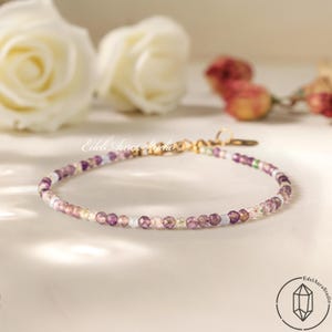 Può includere: Delicato braccialetto di perline con piccole pietre preziose sfaccettate nei toni del viola, rosa e bianco. Il braccialetto ha una chiusura color oro ed è esposto su una superficie chiara. Il braccialetto è di Edel Aura Studio.