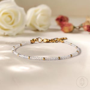 Puede incluir: Delicada pulsera con pequeñas cuentas azul claro y detalles dorados. La pulsera tiene un cierre dorado y se muestra sobre una superficie blanca con elementos florales borrosos en el fondo. El texto "@Edd Aura Studio" es visible.