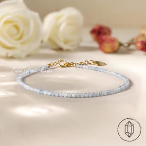 Può includere: Un delicato braccialetto con piccole perline azzurre infilate insieme con una chiusura color oro. Il braccialetto è esposto su una superficie bianca, con rose bianche e altri fiori sullo sfondo. Il braccialetto è di Edel Aura Studio.