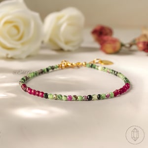 May include: A delicate beaded bracelet featuring a mix of ruby, green, and black gemstones. The bracelet has a gold clasp and is displayed on a white surface with blurred flowers in the background. The bracelet is a jewelry item.
