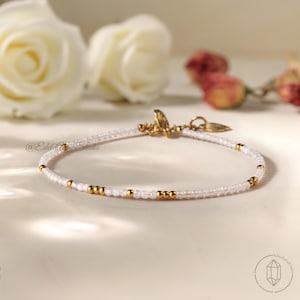 Könnte beinhalten: Zartes Armband mit kleinen, klaren Perlen und goldfarbenen Perlen. Das Armband hat einen goldenen Verschluss und einen kleinen Anhänger. Der Hintergrund umfasst weiße Rosen. Das Armband ist ein Schmuckstück.