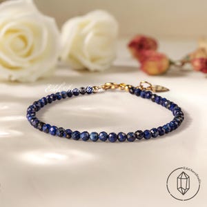 May include: A delicate bracelet featuring small, faceted, deep blue lapis lazuli beads. The bracelet has a gold-colored clasp and a small charm. The background includes white roses and the text "Edel Aura Studio".