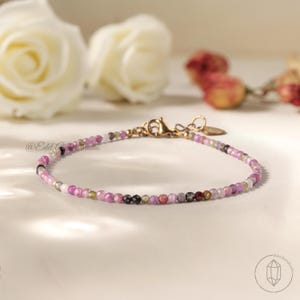 Peut inclure: Bracelet délicat orné de petites pierres précieuses colorées, principalement roses, violettes et vertes. Il est doté d'un fermoir doré. Le bracelet est présenté sur une surface blanche. C'est un bijou.