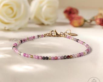 May include: A delicate bracelet with small, multi-coloured gemstones, mainly pink, purple, and green. It has a gold-coloured clasp. The bracelet is displayed on a white surface. The bracelet is a piece of jewellery.