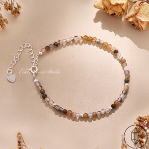 Peut inclure: Un bracelet composé de petites perles rondes multicolores dans des tons marron, gris et blanc translucide, alternées avec de minuscules perles métalliques argentées. Il comprend une chaînette d'extension argentée avec un petit pendentif cœur.