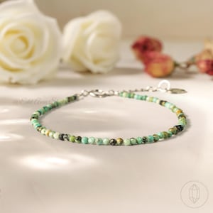 May include: A delicate bracelet featuring small, round turquoise beads strung on a silver chain. The bracelet is displayed on a white surface with blurred white roses and dried flowers in the background. The text "@Edd Aura Studio" is visible.