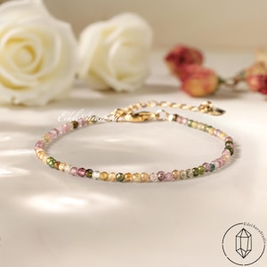 Può includere: Braccialetto delicato con piccole perline di pietre preziose multicolori. Il braccialetto ha una catena e una chiusura color oro. Lo sfondo include rose bianche e un logo "Edel Aura Studio".