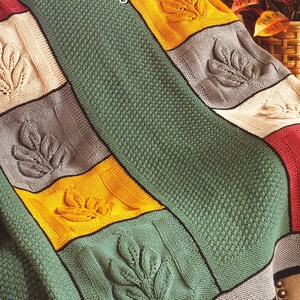 May include: A knitted blanket with a patchwork design featuring leaf motifs. The blanket has a green background with squares in beige, gray, yellow, and red. Each square has a raised leaf design, and the edges are outlined in black.