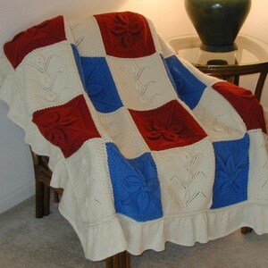 May include: A white knitted throw blanket with a red, white, and blue patchwork design. The blanket features a floral pattern and is draped over a chair.