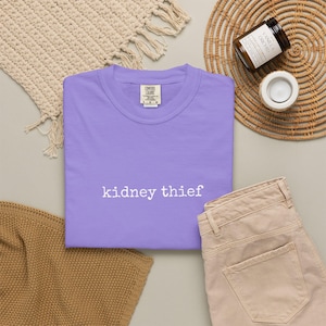 May include: A light purple short-sleeved t-shirt with a crew neckline. The shirt features the text "kidney thief" printed in white, lowercase, typewriter-style font across the center of the chest. A Comfort Colors brand label is visible at the neck.