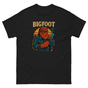 Puede incluir: Camiseta negra con un gráfico de Bigfoot. La ilustración muestra a Bigfoot con gafas y una camisa a cuadros, sosteniendo una taza con vapor. La palabra "BIGFOOT" está impresa encima del gráfico en negrita, en naranja.