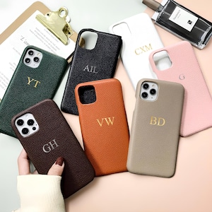 Puede incluir: Una colección de fundas de teléfono personalizadas en varios colores, incluyendo verde oscuro, negro, marrón, naranja, blanco, rosa y beige. Cada funda presenta iniciales doradas. También se ve una carpeta con texto y un frasco de perfume.