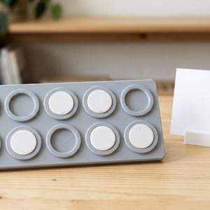 May include: A rectangular grey ten-frame tray featuring ten circular recessed slots, with seven white circular discs placed inside the slots. Beside the tray stands a small white rectangular card holder containing a blank white rectangular card.