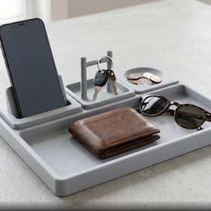 May include: A light gray desk organizer tray featuring a dedicated upright slot for a smartphone, a small square compartment with a central post for hanging keys, a circular dish for loose coins, and a large rectangular area for a wallet and sunglasses.