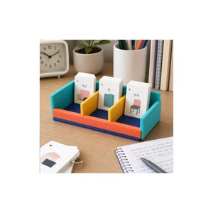 Puede incluir: Un organizador de escritorio rectangular con laterales color verde azulado, un panel frontal naranja y una base azul oscuro. Cuenta con dos divisores amarillos que crean tres compartimentos, cada uno con una tarjeta con texto coreano e ilustraciones.