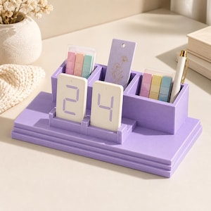 May include: A lavender desk organiser with three compartments, holding two white digital calendar blocks displaying the number 24, two sets of pastel sticky notes, a purple floral bookmark, and a white pen with gold accents.
